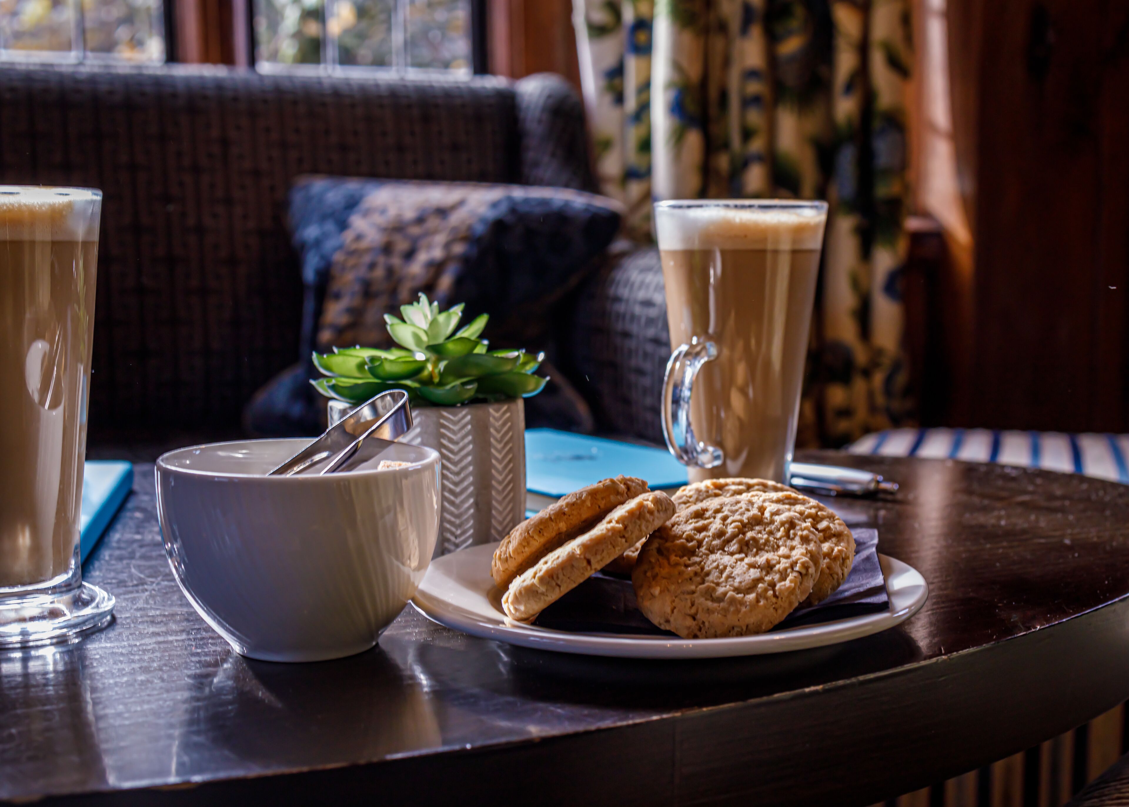 Una mesa con dos tazas de café, un bol con una cuchara, un plato de galletas y una planta en maceta.