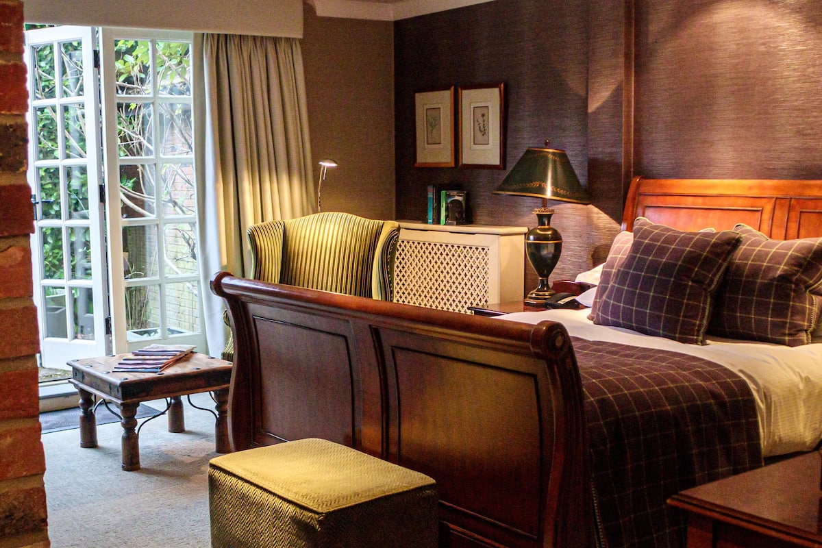 A hotel room with a bed, a chair, a table, and a lamp. There is a window with curtains and a view of greenery outside.