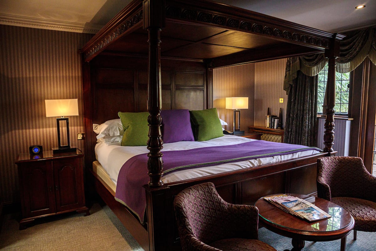 A four-poster bed with a wooden frame, two nightstands with lamps, and two upholstered chairs.
