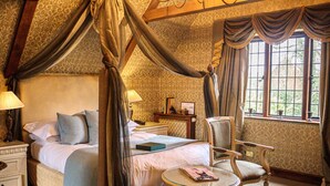 A four-poster bed with a canopy, a bedside table with a lamp, a chair, and a window with curtains. - Langshott Manor (Horley)
