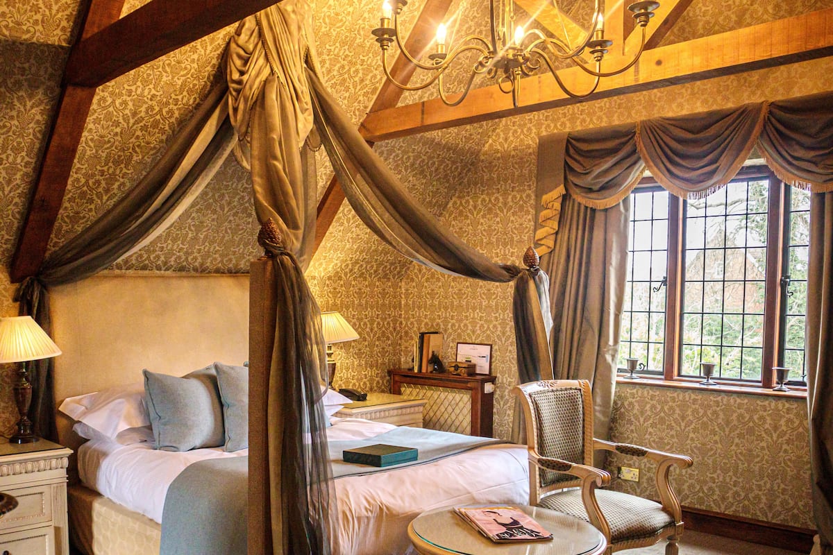 A four-poster bed with a canopy, a bedside table with a lamp, a chair, and a window with curtains.