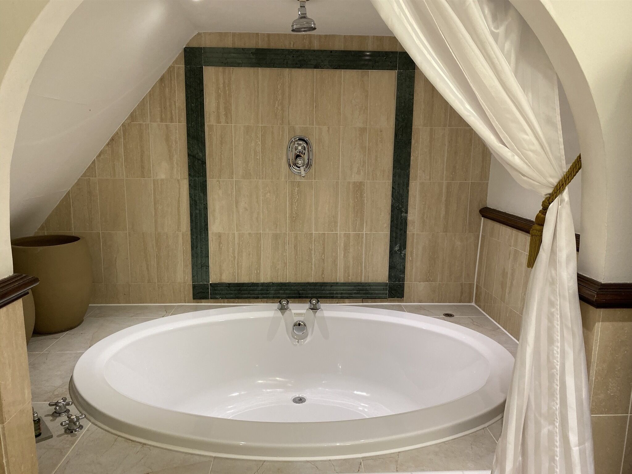 Una bañera ovalada grande en un rincón, con una ducha encima, rodeada de baldosas color beige y una pared de acento verde.