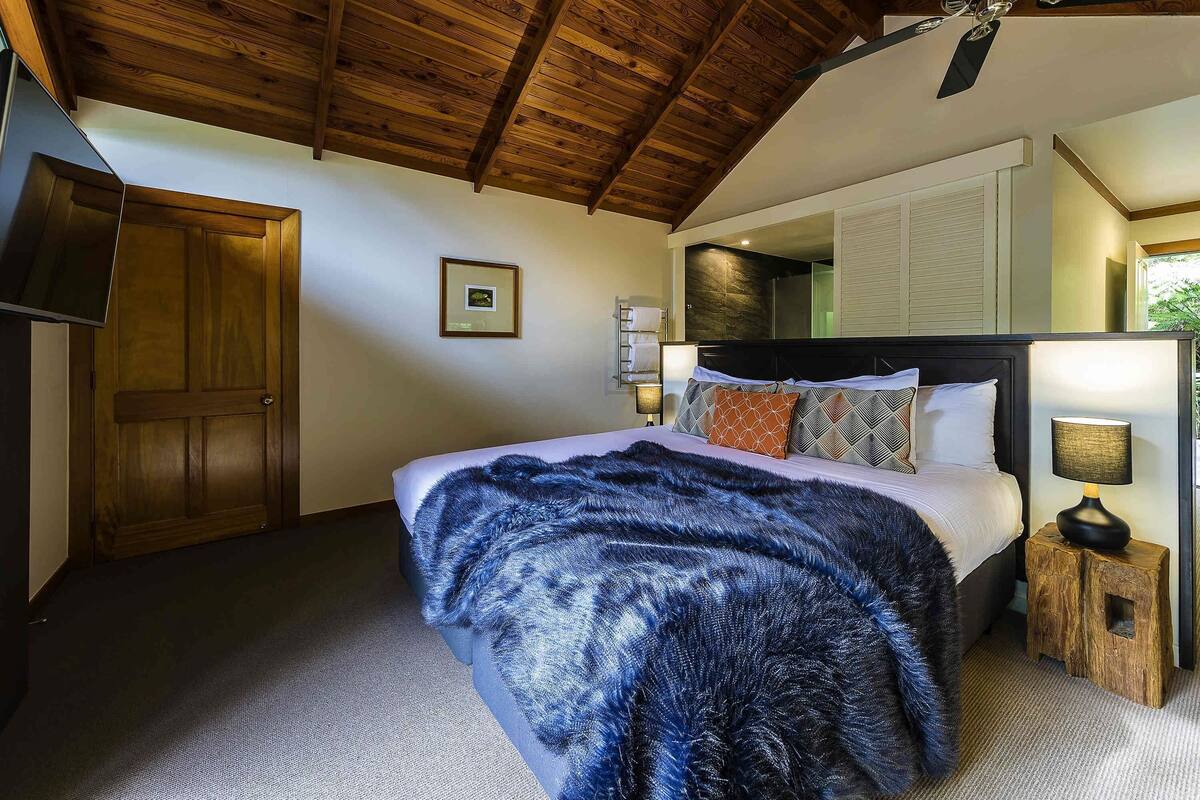 Un dormitorio con techo de madera, cama con almohadas estampadas, mesita de noche con lámpara y una puerta.