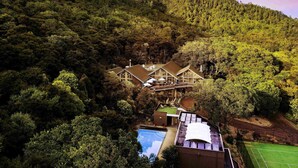 Une grande maison entourée d’une forêt dense, avec une piscine et un court de tennis visibles.