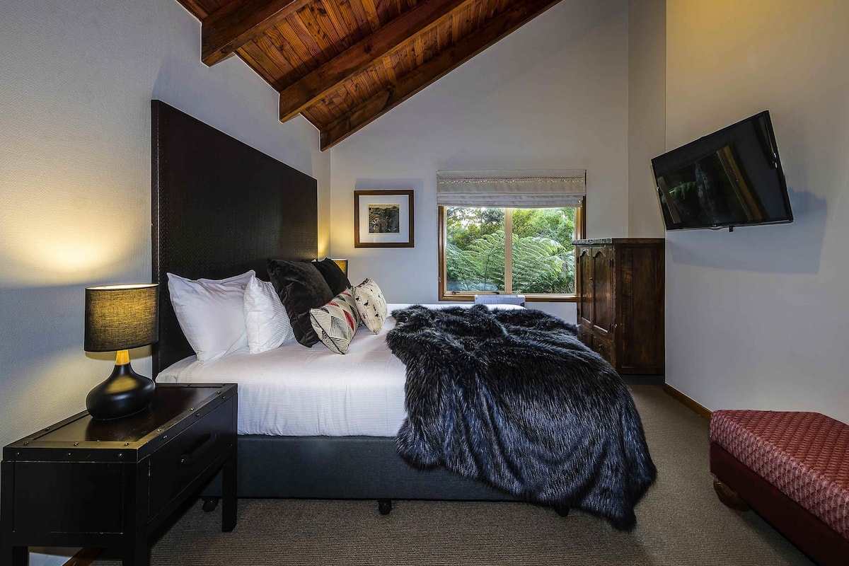 Un dormitorio con cama, mesita de noche, lámpara, televisor y ventana con vista a árboles.