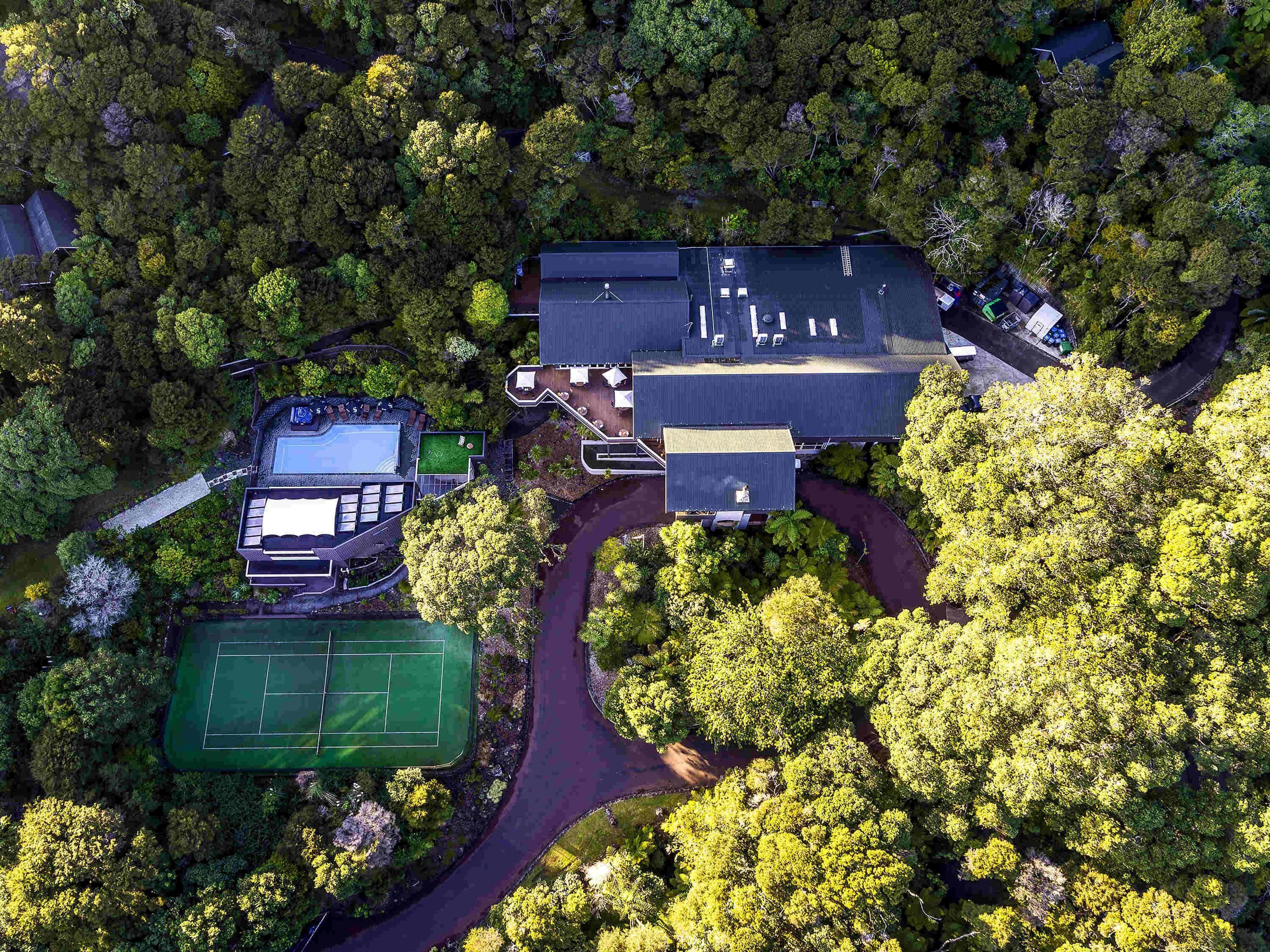 An aerial view of a residential property with a swimming pool, tennis court, and a large house surrounded by dense trees.