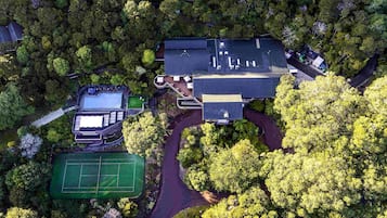 An aerial view of a residential property with a swimming pool, tennis court, and a large house surrounded by dense trees.
