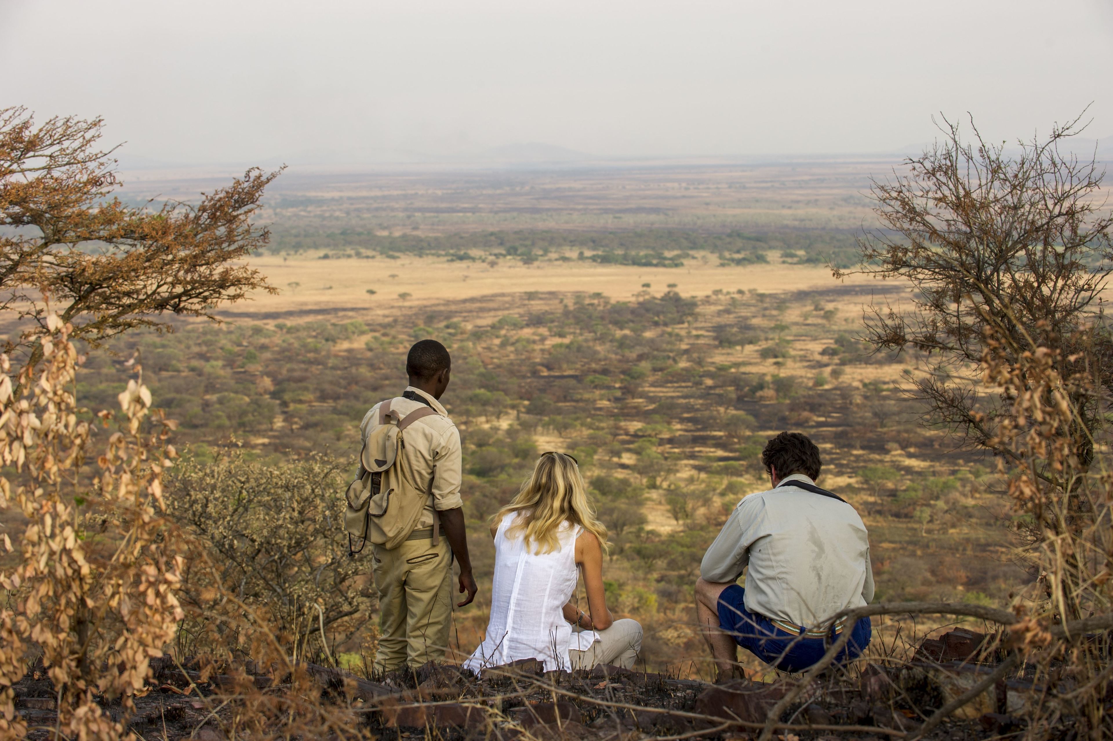 Three people sitting on a rocky outcrop overlooking a vast savannah landscape.