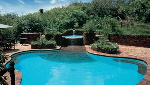 A swimming pool with a waterfall feature, surrounded by lush greenery and a clear blue sky. - Kirawira Serena Camp (Serengeti)