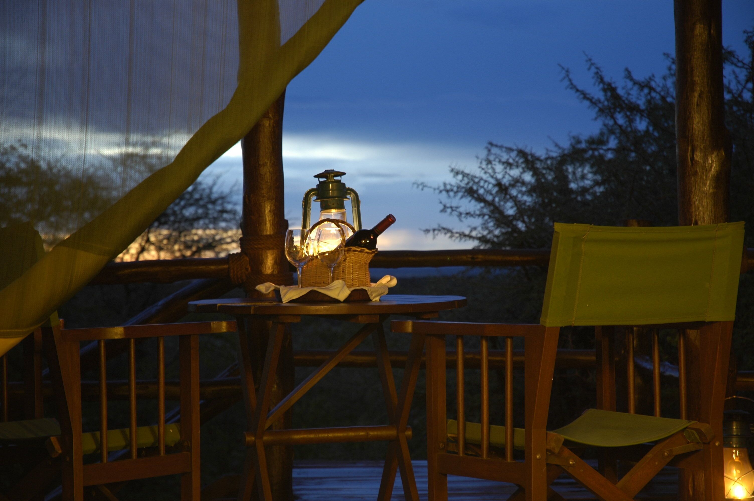 A wooden deck with a table set for dining, a lantern, and a bottle of wine.