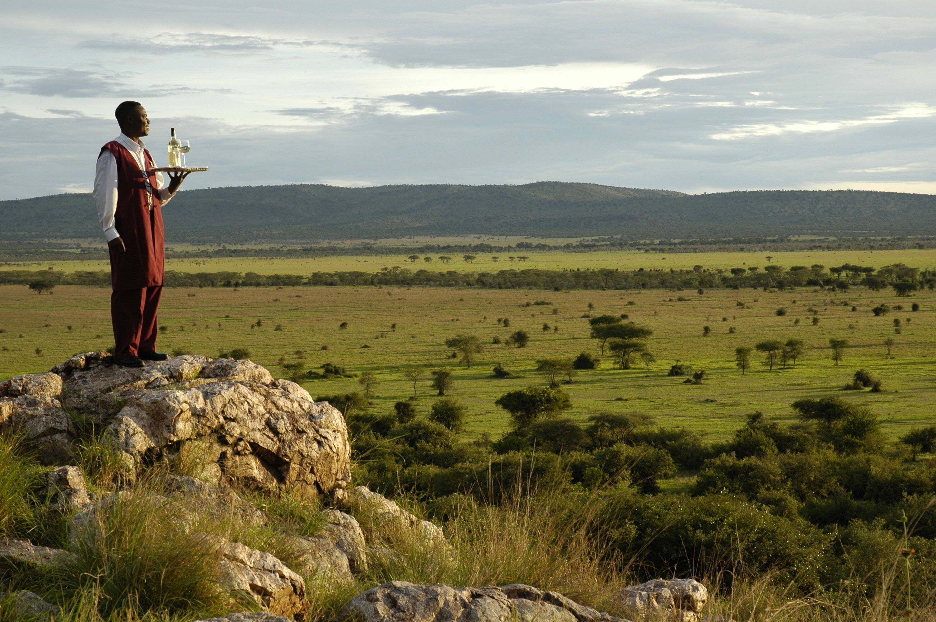 A person standing on a rock with a tray of bottles, overlooking a vast landscape with trees and hills.