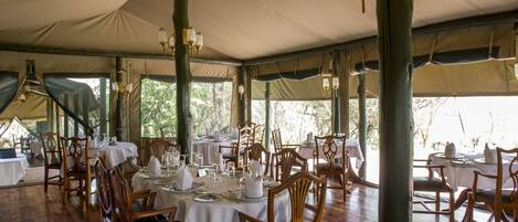 A tented dining area with tables set for service, wooden chairs, and hanging light fixtures.