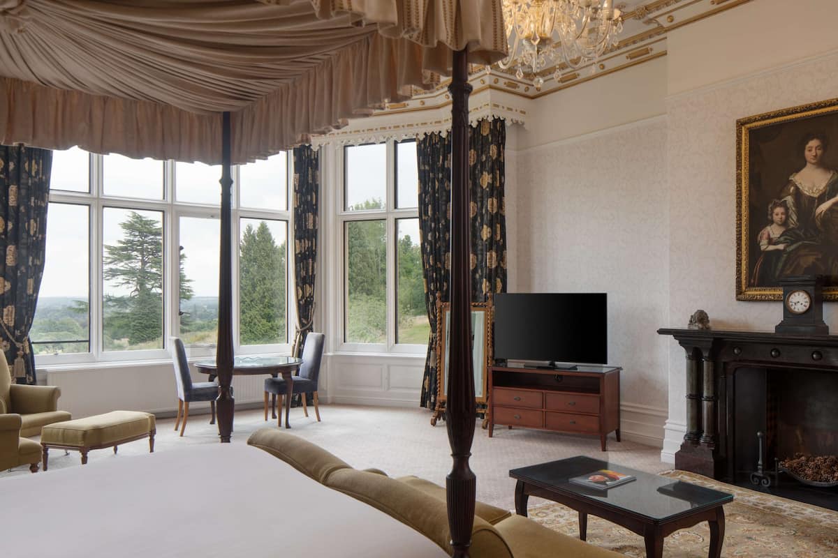 A spacious room with a large bed, a television, and a fireplace. There are multiple windows offering a view of the outdoors.