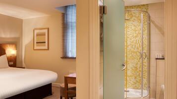 A hotel room with a bed, a desk, a chair, a shower with mosaic tiles, and a window with blinds.