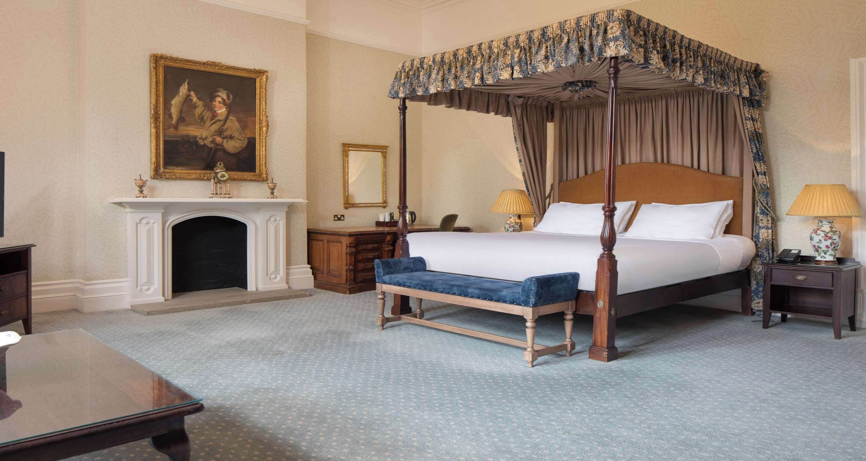 A four-poster canopy bed with a blue upholstered bench, a fireplace, and a painting on the wall.
