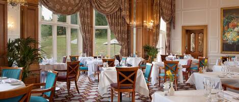 A formal dining room with multiple tables set for service, large windows with curtains, and a chandelier.