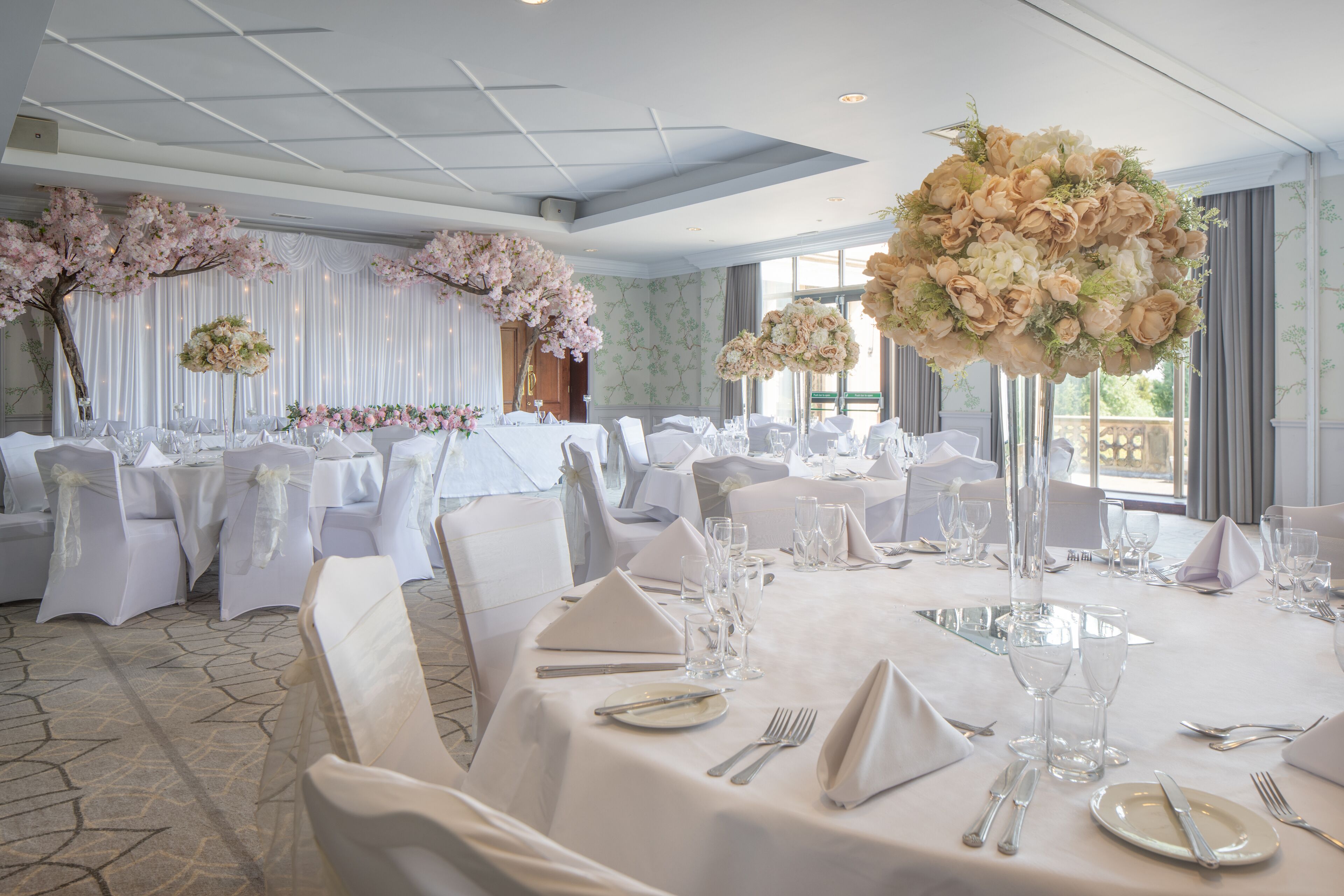 Un salón de banquetes con mesas redondas dispuestas para un evento formal, decorado con centros de mesa florales y manteles blancos.