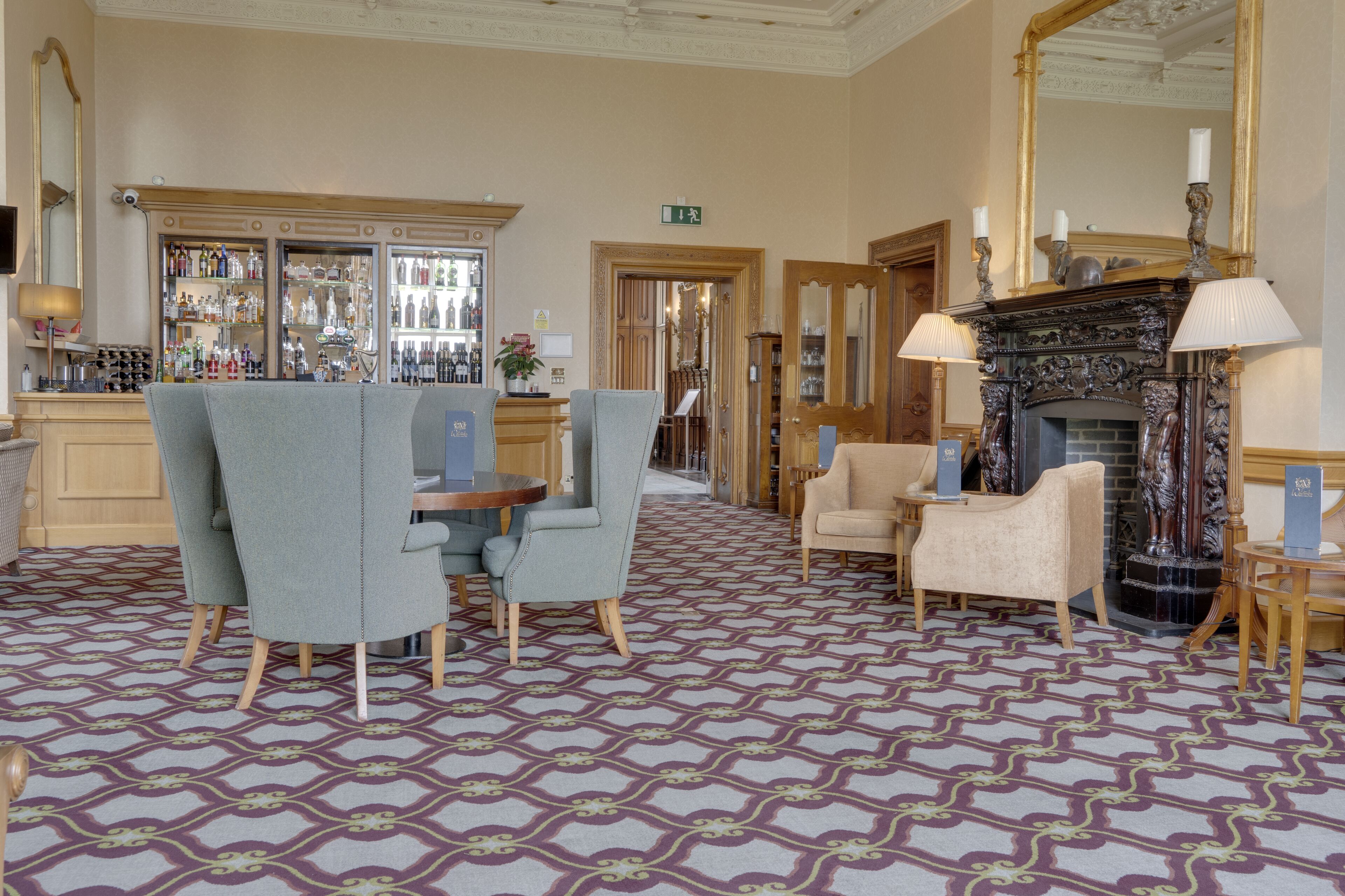 Een hotel lobby met een kamerbreed tapijt, zithoeken, een bar met flessen en een open haard.