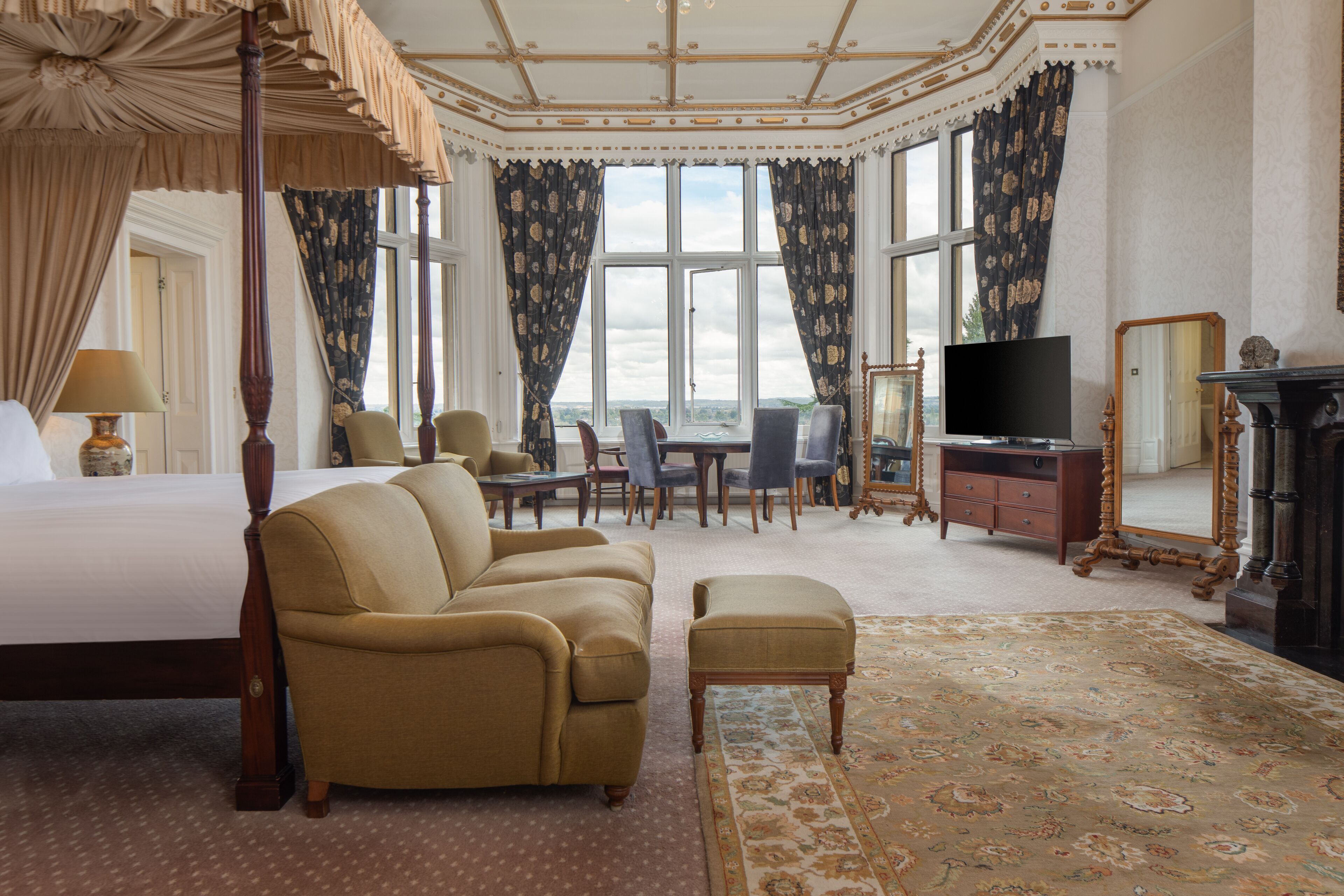 A hotel room with a four-poster bed, a sofa, an ottoman, a desk with chairs, a television, a fireplace, and large windows with curtains.