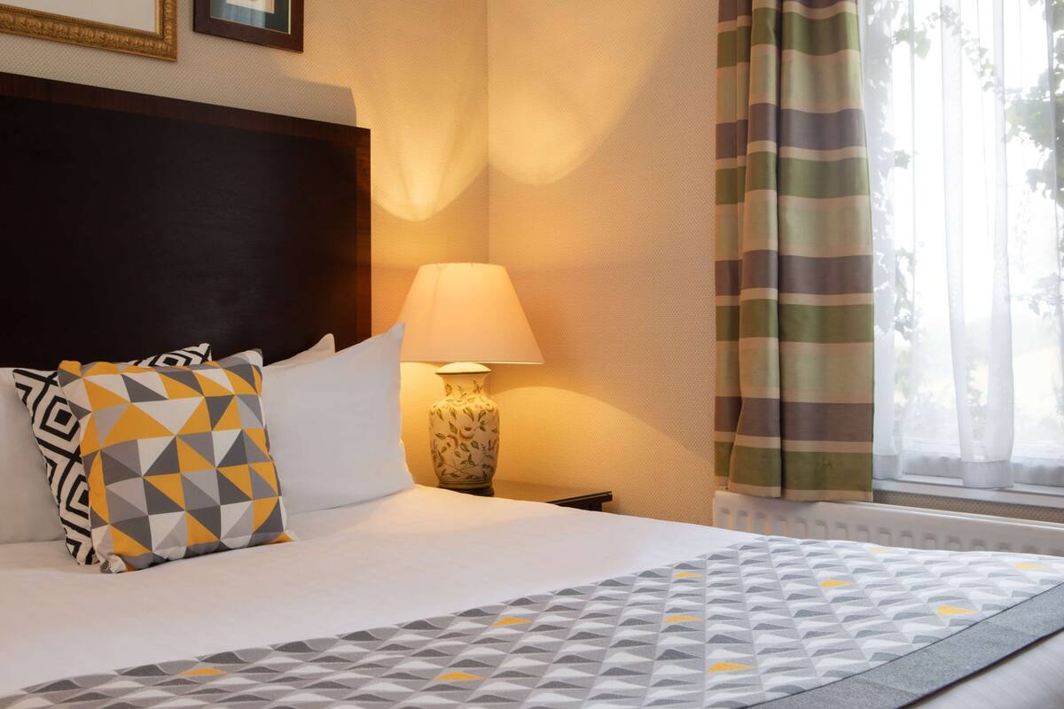 A neatly made bed with patterned pillows, a bedside lamp, and a window with curtains.