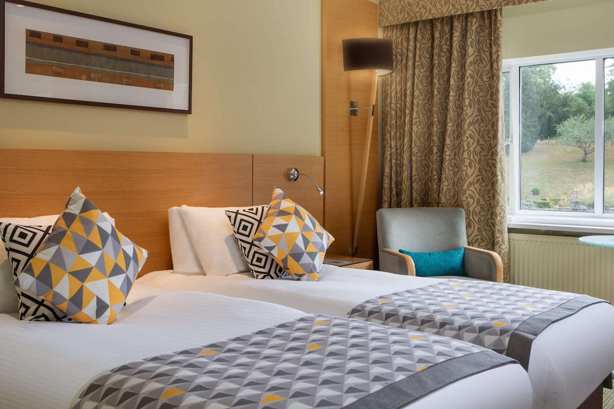 A hotel room with two beds, a chair, a small table, a window with a view of greenery, and a framed picture on the wall.