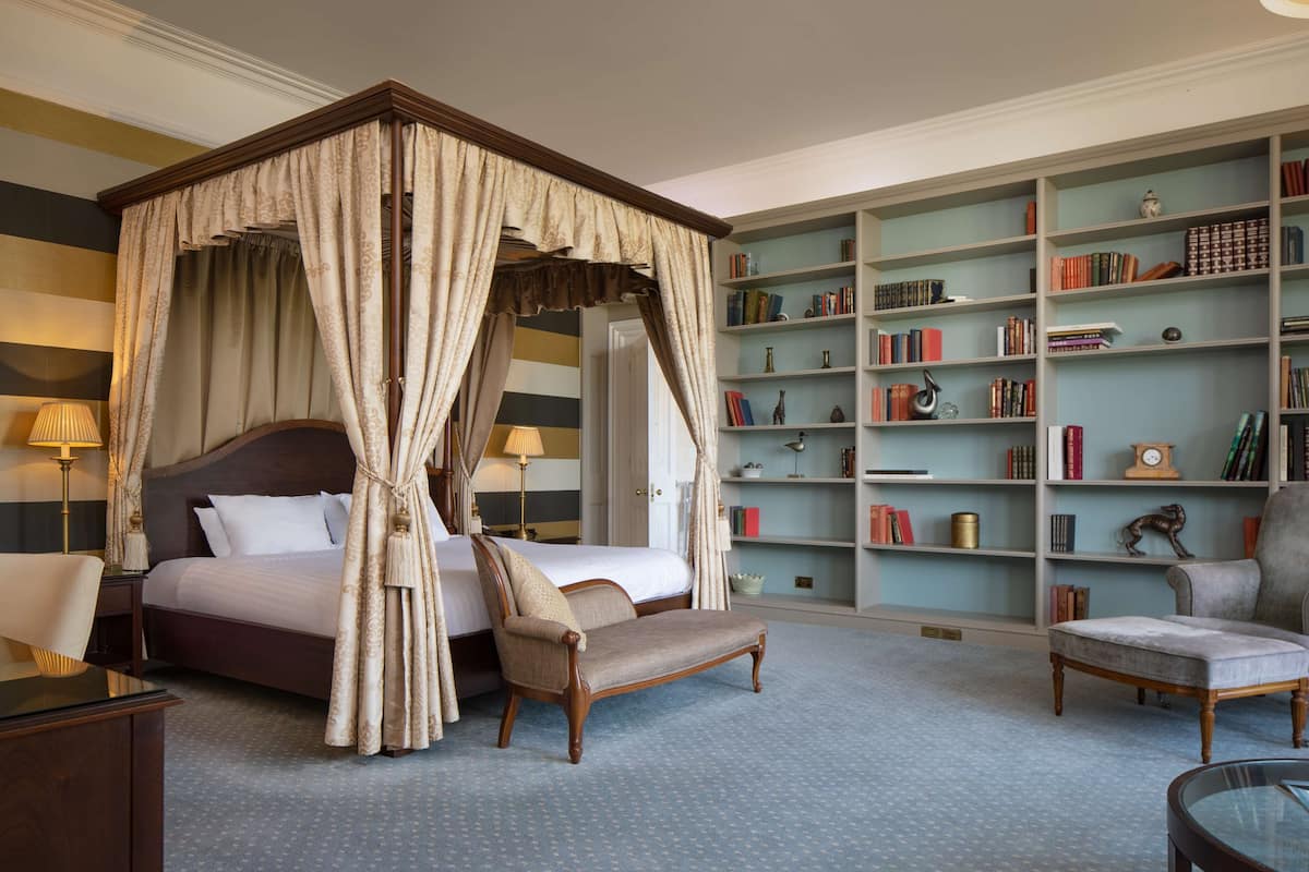 A four-poster canopy bed with a nightstand, a chair, a bookshelf, and a round table in a hotel room.