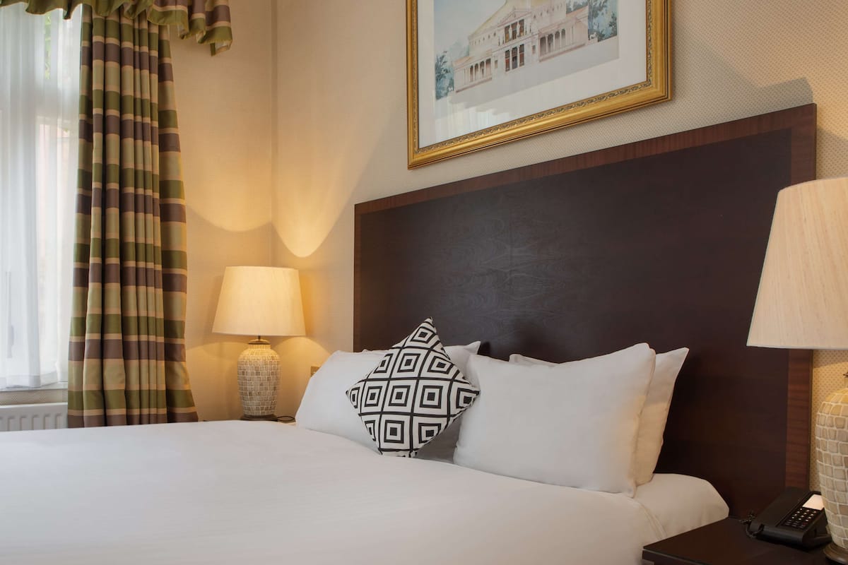 A hotel room with a large bed, a headboard, a bedside table with a lamp, a telephone, and a window with curtains.