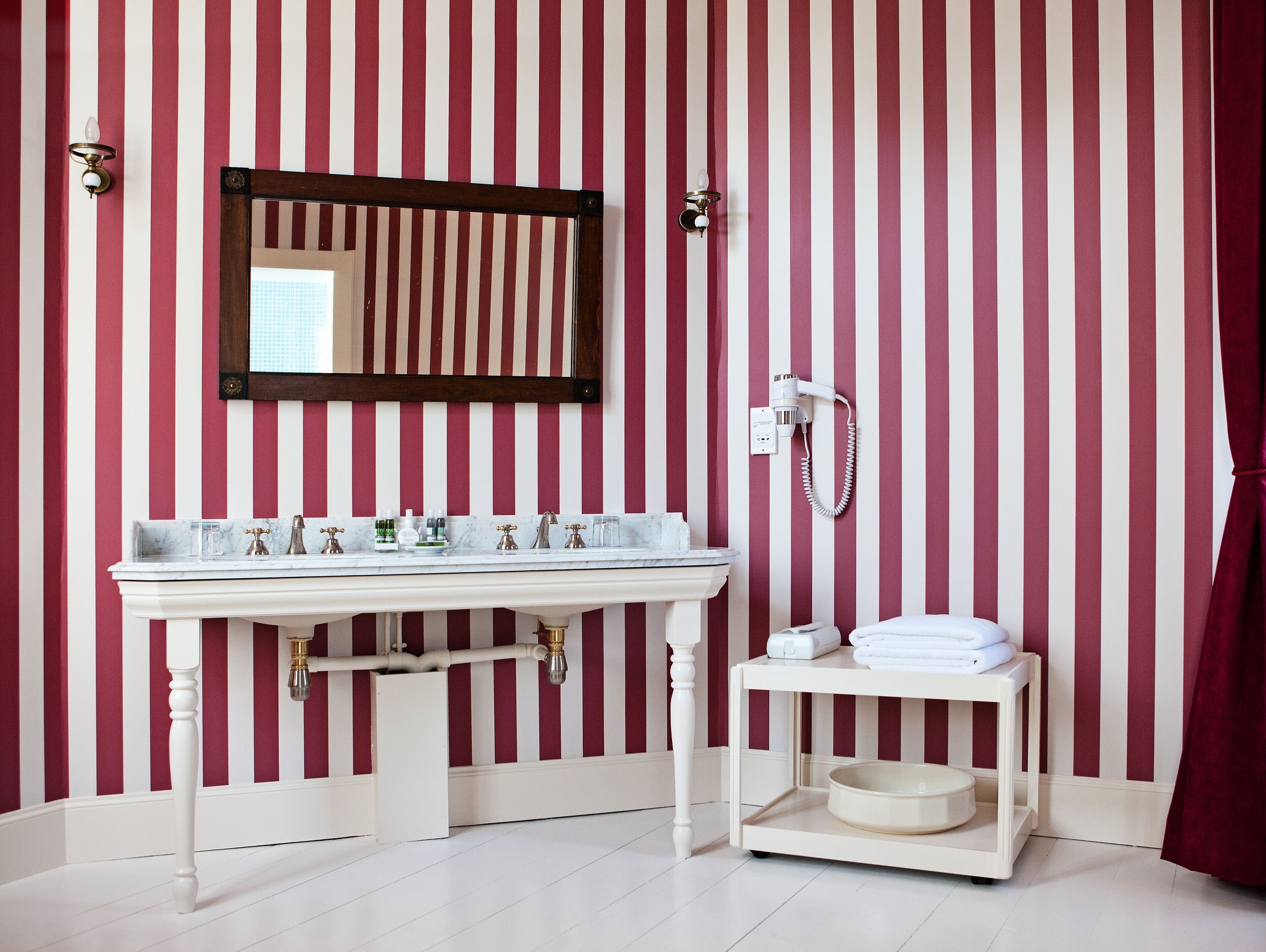A bathroom with a striped red and white wall, a white sink with multiple faucets, a mirror, a hairdryer, and a small shelf with towels.