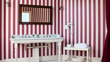 A bathroom with a striped red and white wall, a white sink with multiple faucets, a mirror, a hairdryer, and a small shelf with towels.