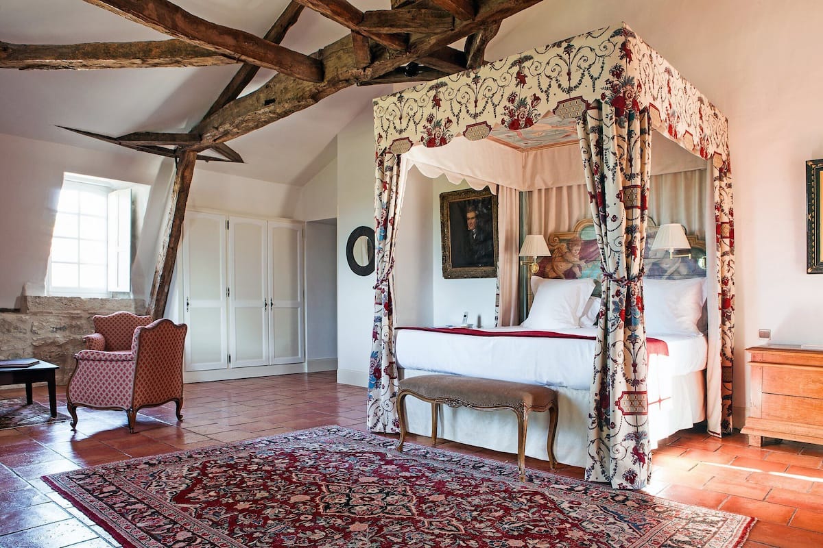 A bedroom with a four-poster canopy bed, a bench, a chair, and a large rug.