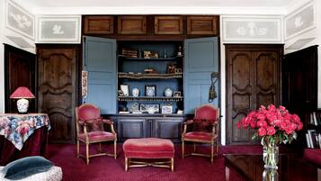 Une pièce avec un tapis rouge, deux fauteuils à revêtement rouge, un pouf en velours rouge, un meuble en bois avec des étagères et un vase rempli de fleurs rouges.