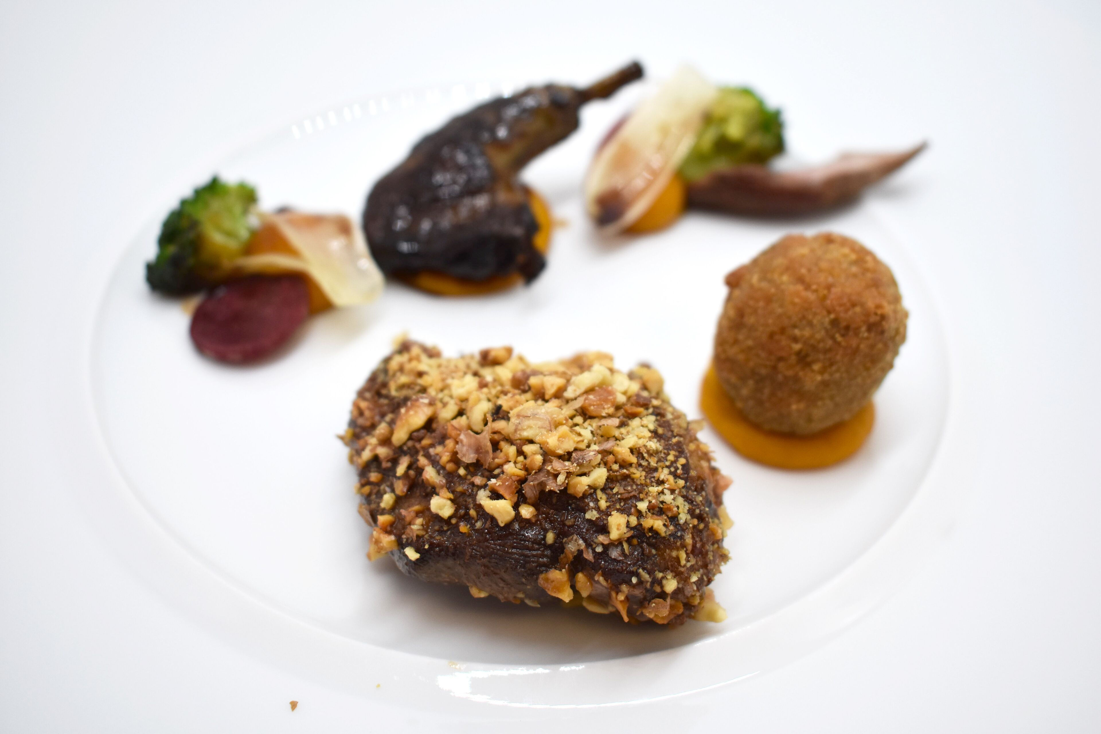 A plate of food with a piece of meat topped with nuts, a round breaded item, and various vegetables.
