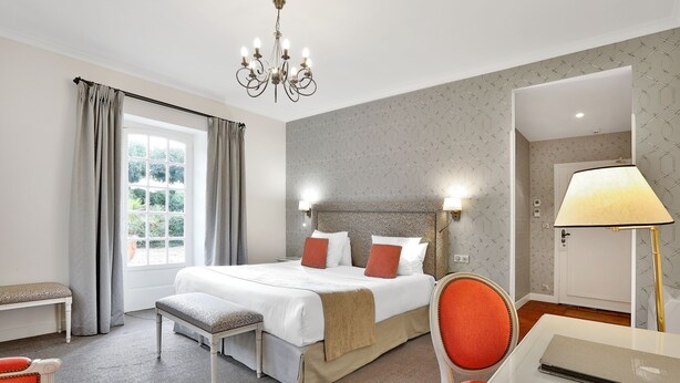 Une chambre d’hôtel avec un grand lit, une banquette, un bureau avec une lampe et une vue sur la verdure par la fenêtre.