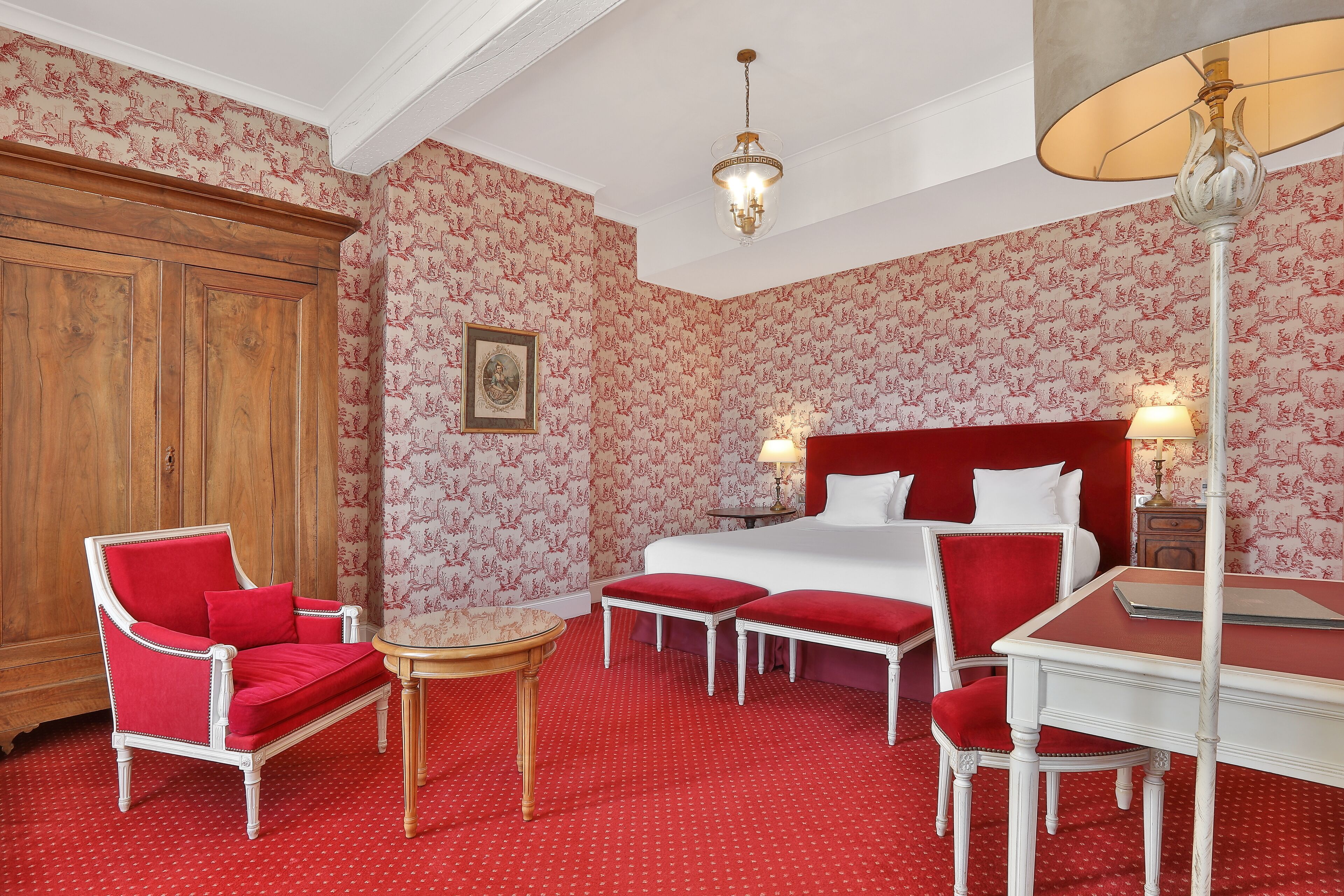 Habitación de hotel con alfombra roja, armario de madera, sillón rojo, una pequeña mesa redonda, cama con ropa blanca y una butaca tapizada en rojo.