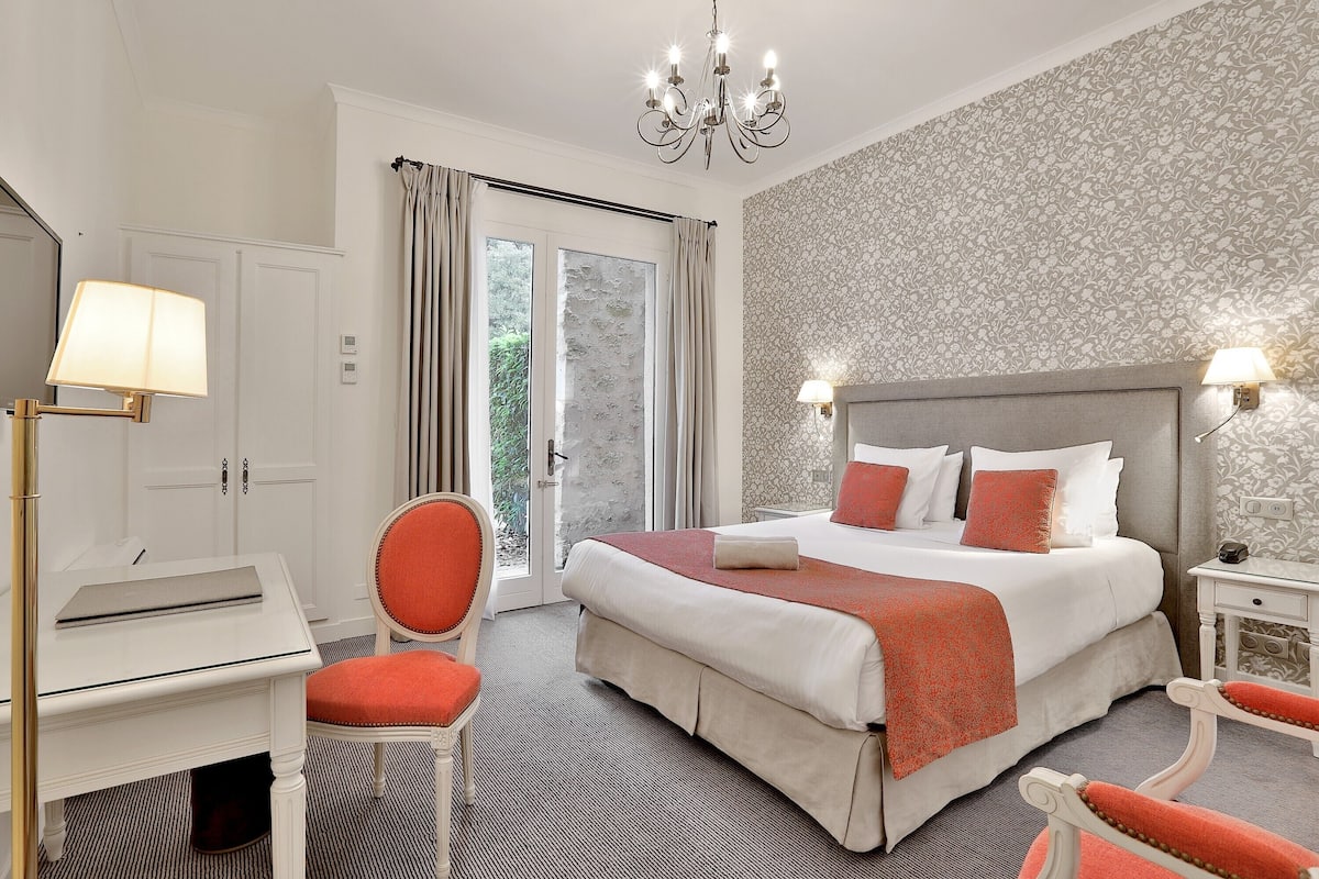 A hotel room with a bed, a desk, a chair, a chandelier, and a view of the outdoors.
