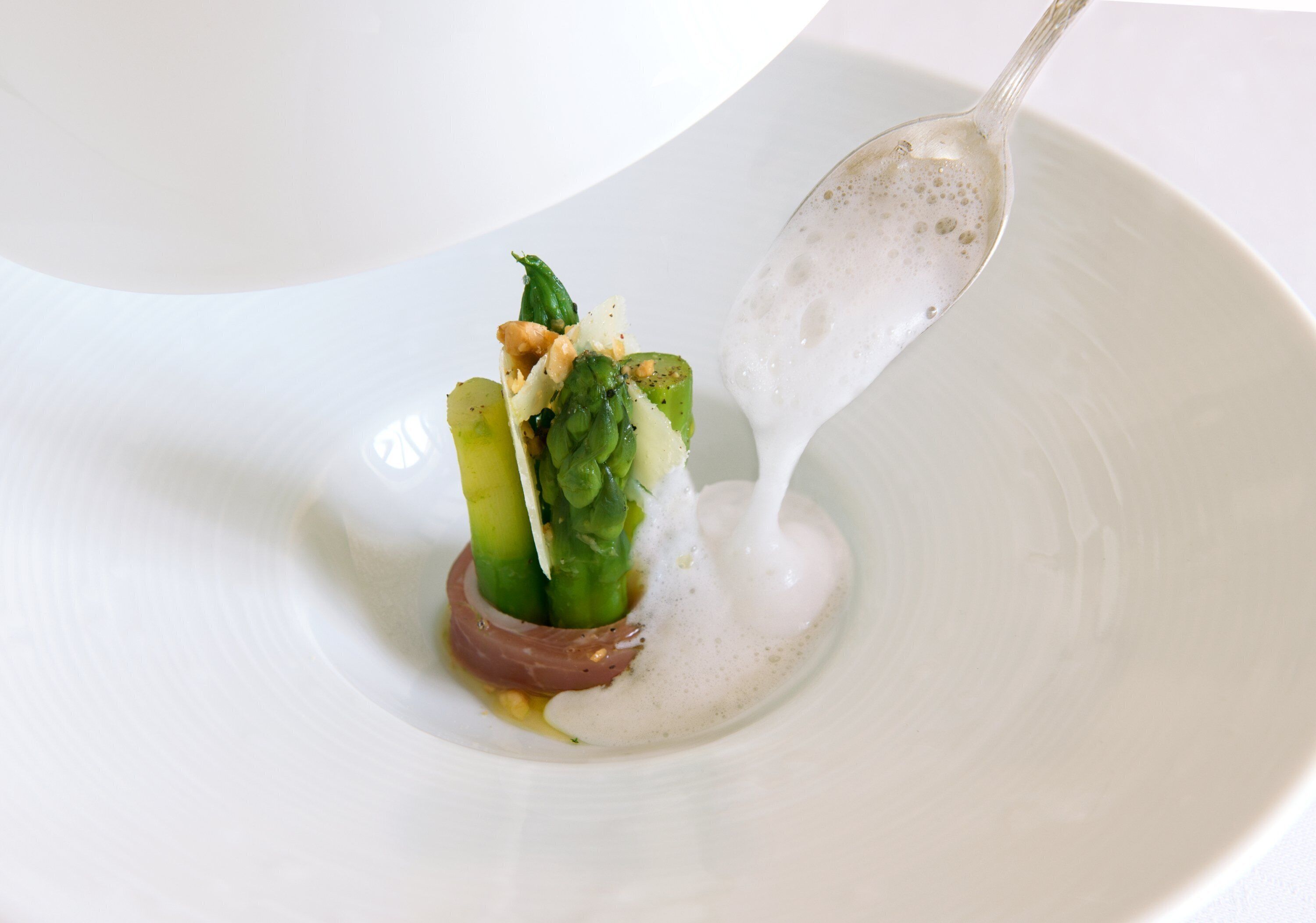 Un plato blanco con una cucharada de salsa cremosa de color blanco vertiéndose sobre un plato que contiene espárragos y una porción de carne.