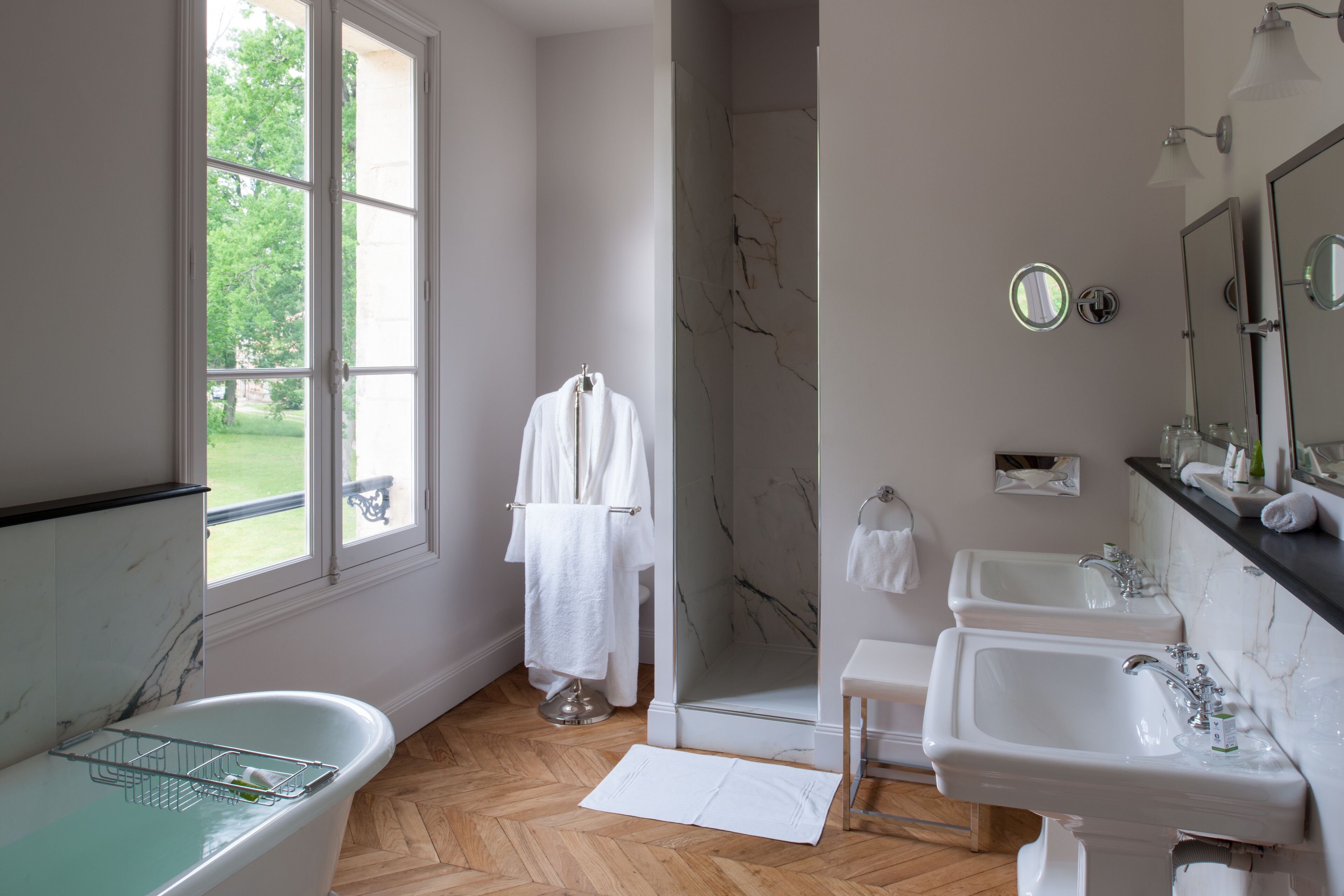 A bathroom with a bathtub, two sinks, a shower, and a window with a view of greenery.