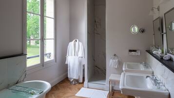 A bathroom with a bathtub, two sinks, a shower, and a window with a view of greenery.