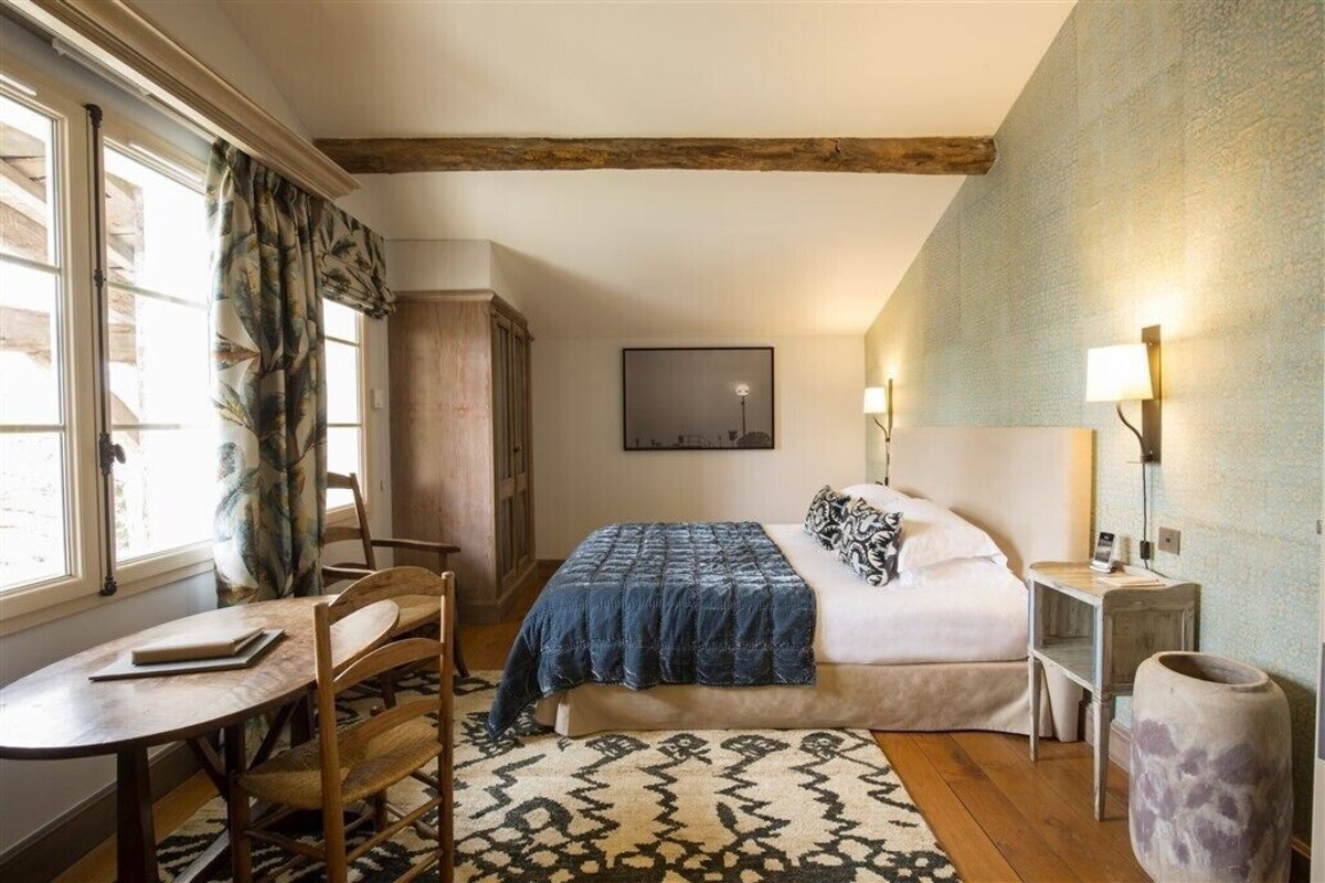 Ein Schlafzimmer mit Bett, Nachttisch, Lampe, Sessel, Schreibtisch und einem Fenster mit Vorhängen.