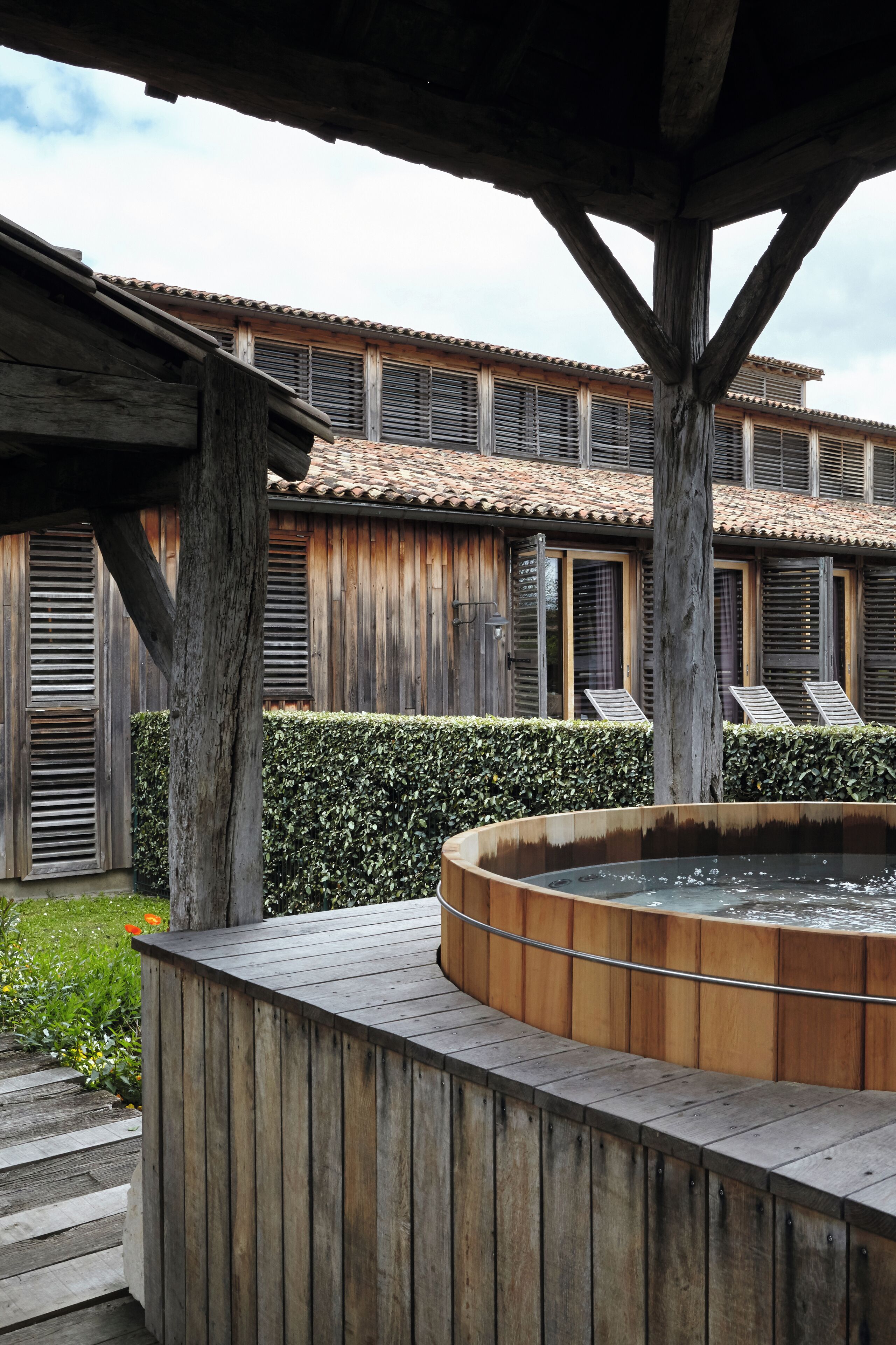 A wooden hot tub under a wooden pergola with a tiled roof building in the background.