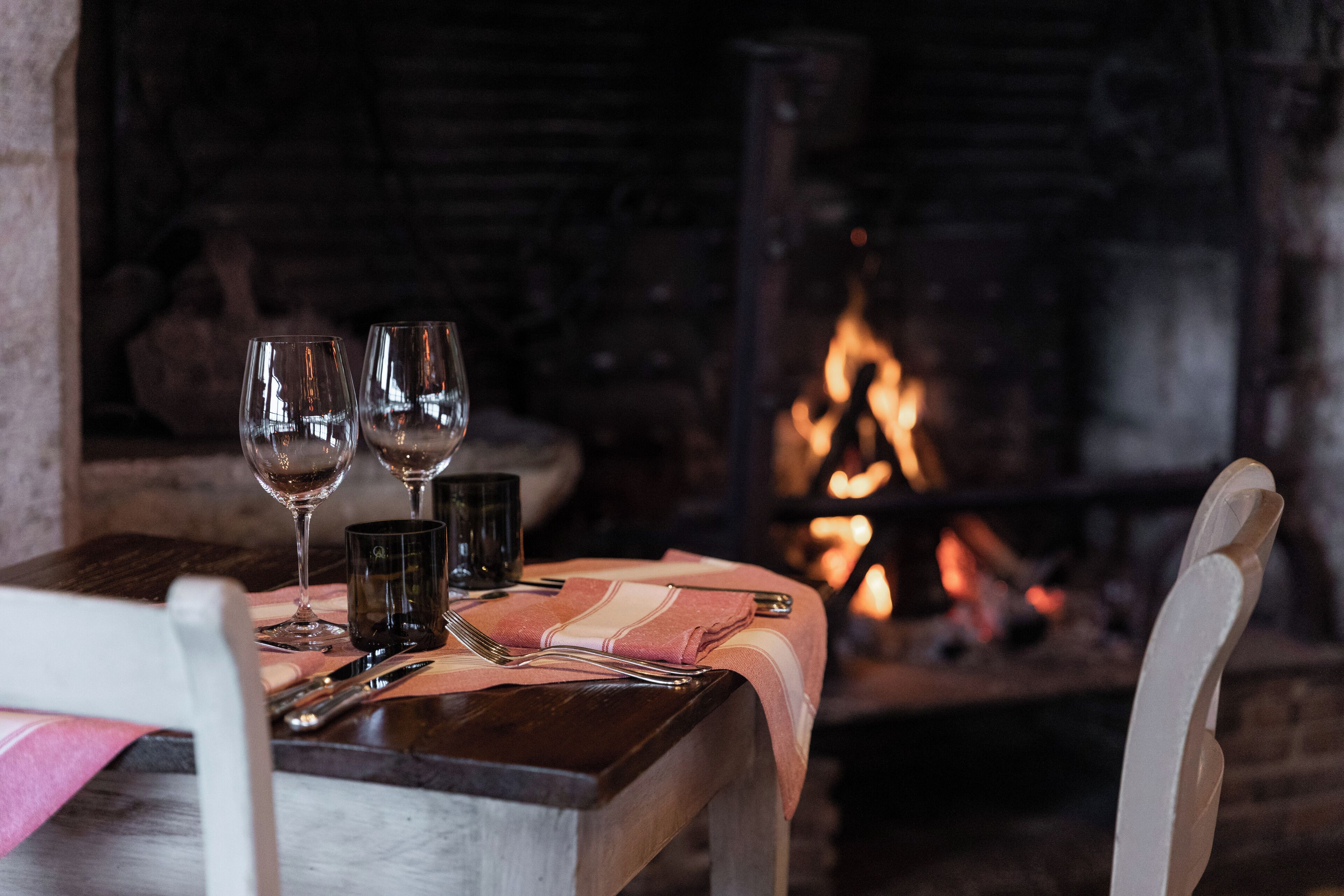 Mesa servida con copas de vino, cubiertos y una vela en una habitación acogedora con una chimenea al fondo.