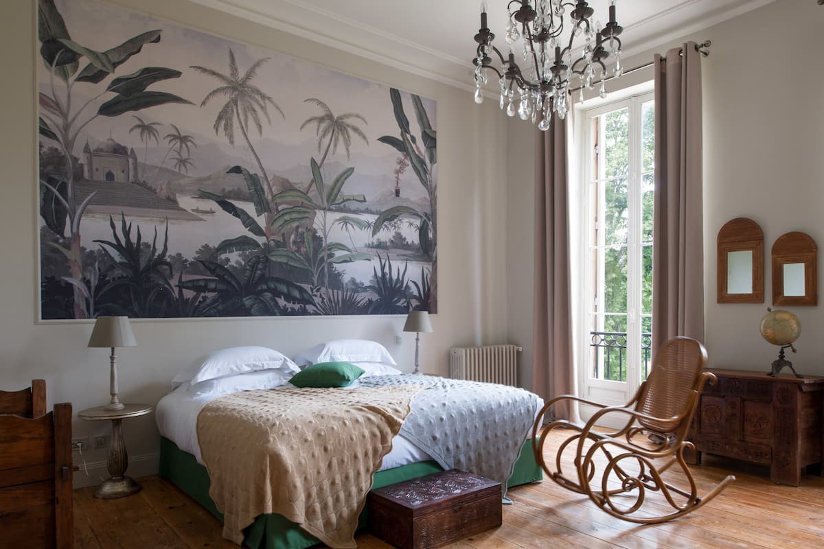 Une chambre avec une grande fresque murale aux motifs tropicaux, un fauteuil à bascule en bois, une table de chevet avec une lampe et une commode surmontée d’un globe terrestre.