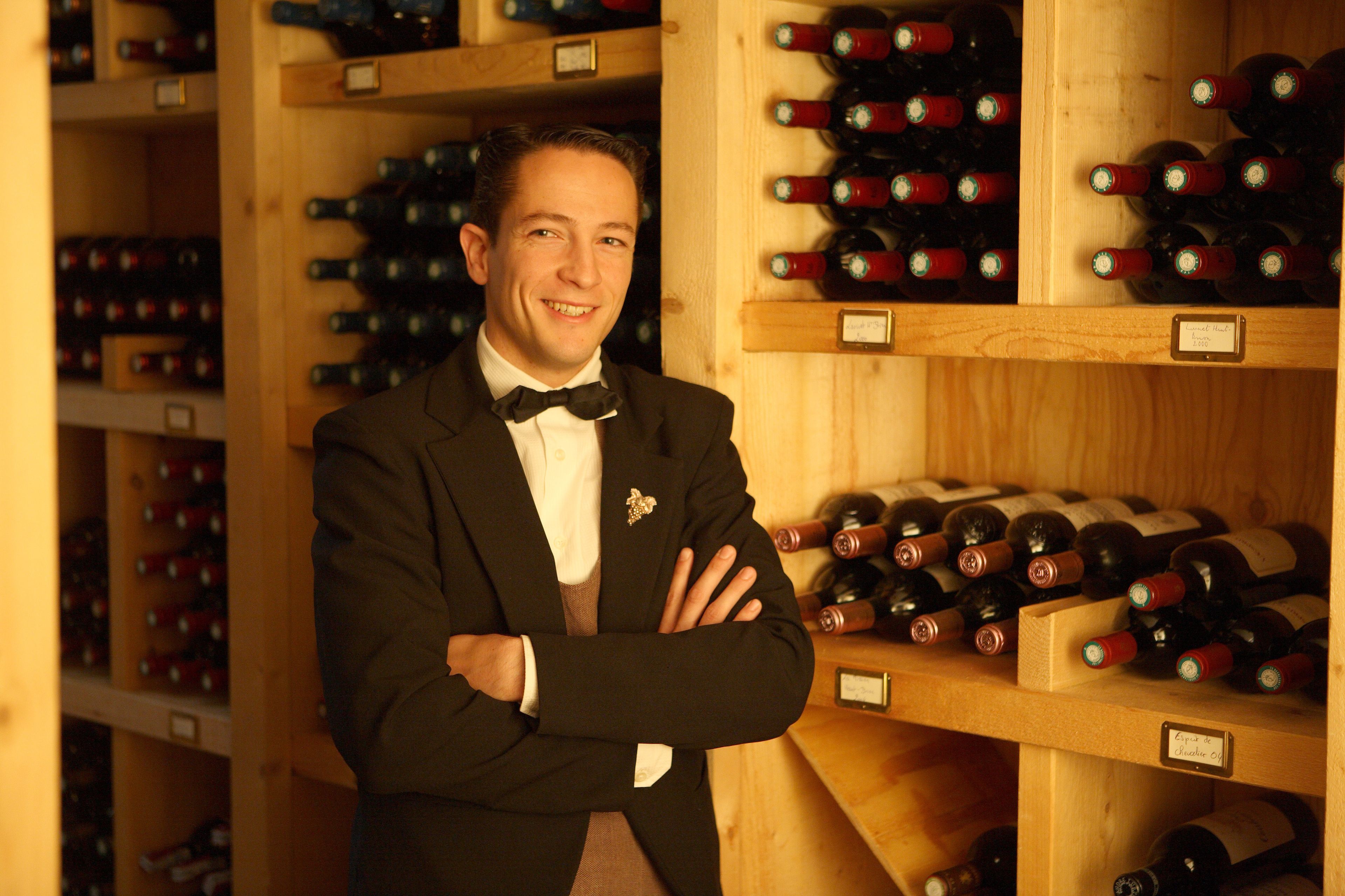 A man in a suit standing in a wine cellar with wooden shelves filled with wine bottles.