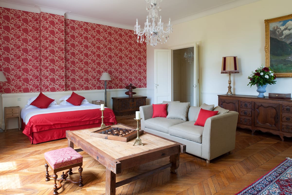 Une pièce comprenant un lit, un canapé, une table basse en bois, un mur décoré d’un motif rouge, un lustre et un tableau accroché au mur.