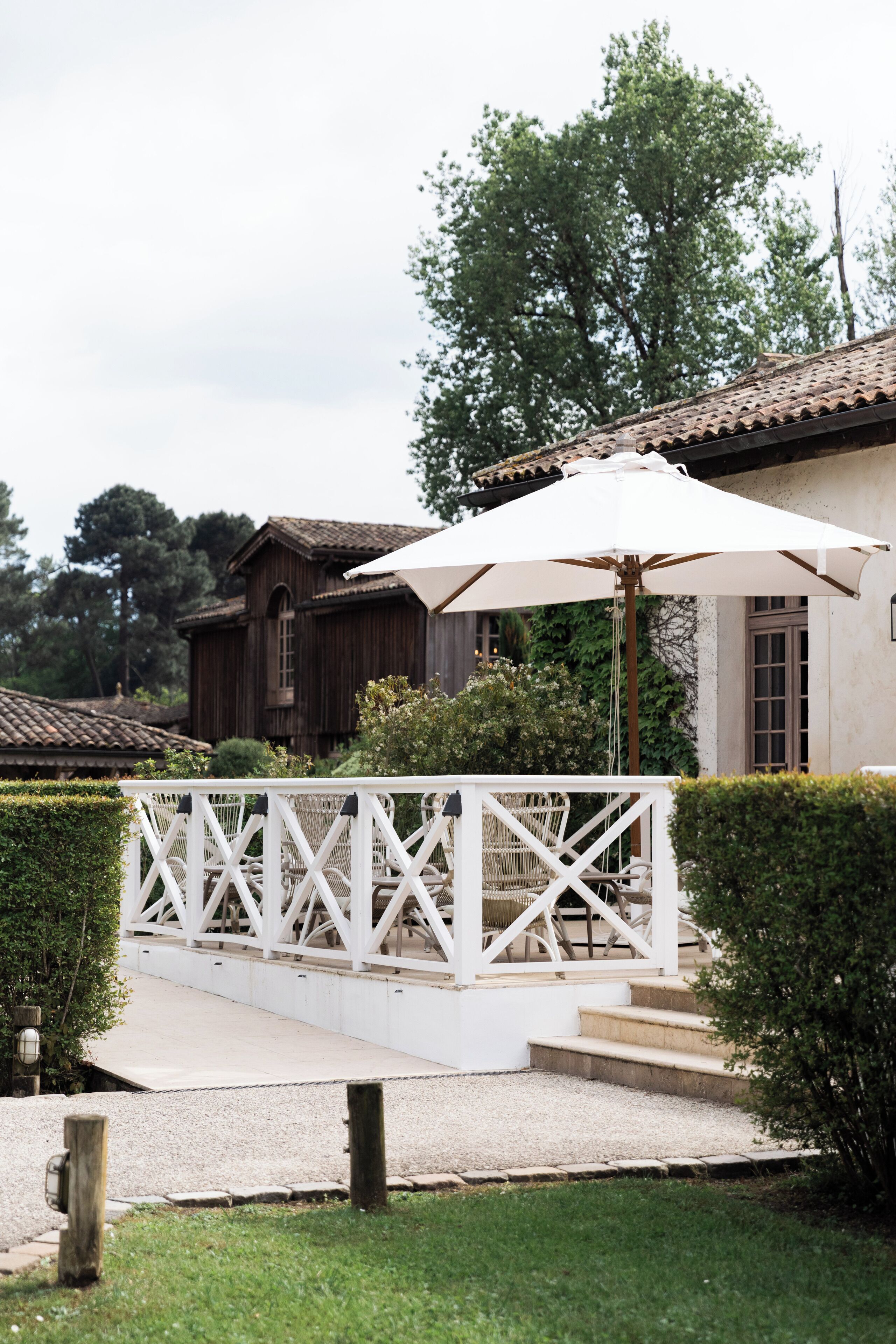 Un área de estar al aire libre con mobiliario blanco, una pérgola y una sombrilla, un edificio de madera al fondo y un seto cuidado en primer plano.