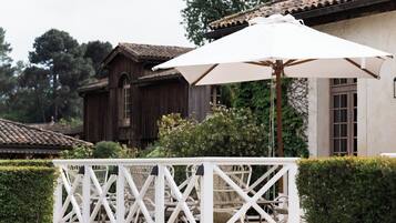 Un área de estar al aire libre con mobiliario blanco, una pérgola y una sombrilla, un edificio de madera al fondo y un seto cuidado en primer plano.