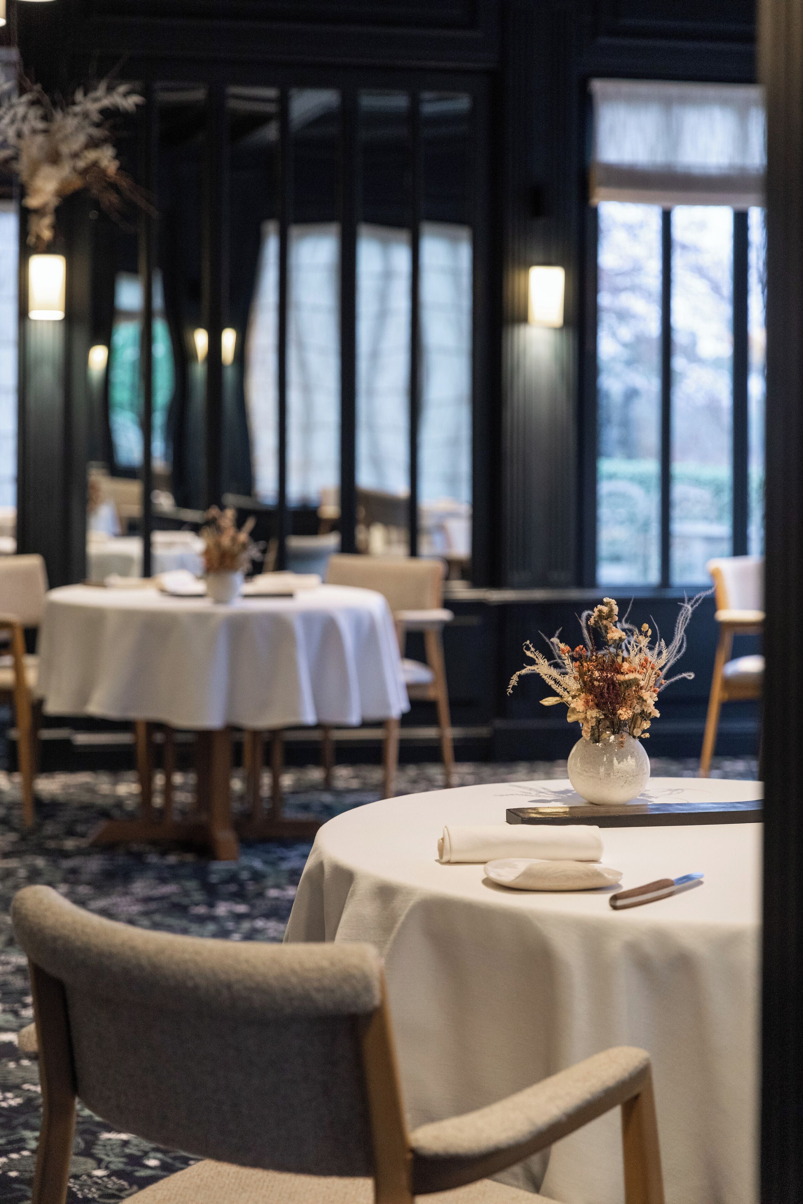 Un restaurante con mesas redondas, manteles blancos y un jarrón de flores secas sobre la mesa.