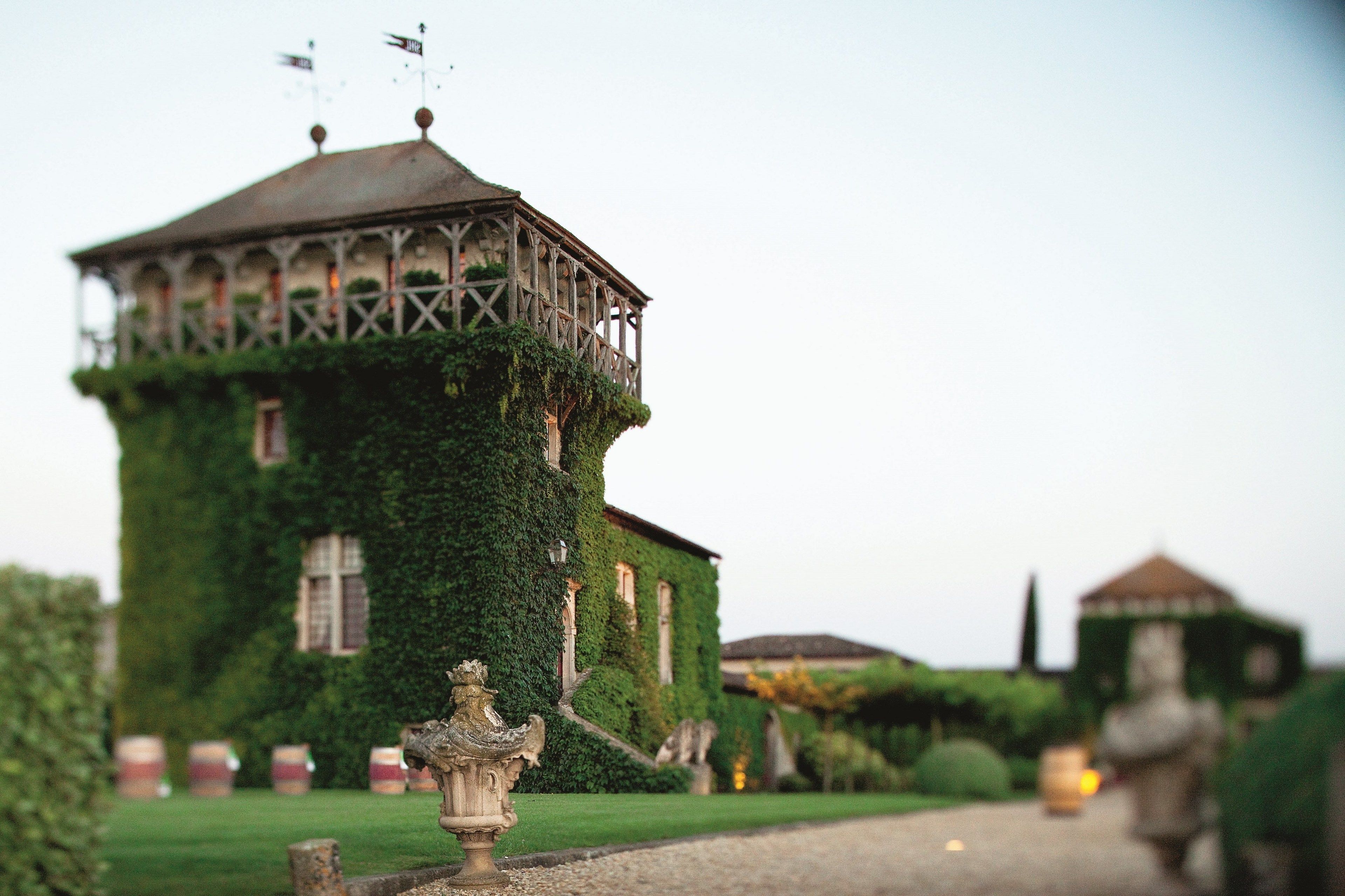 Um edifício com uma torre coberta de hera, uma estátua em primeiro plano e um jardim bem cuidado.