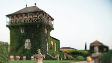 Um edifício com uma torre coberta de hera, uma estátua em primeiro plano e um jardim bem cuidado.