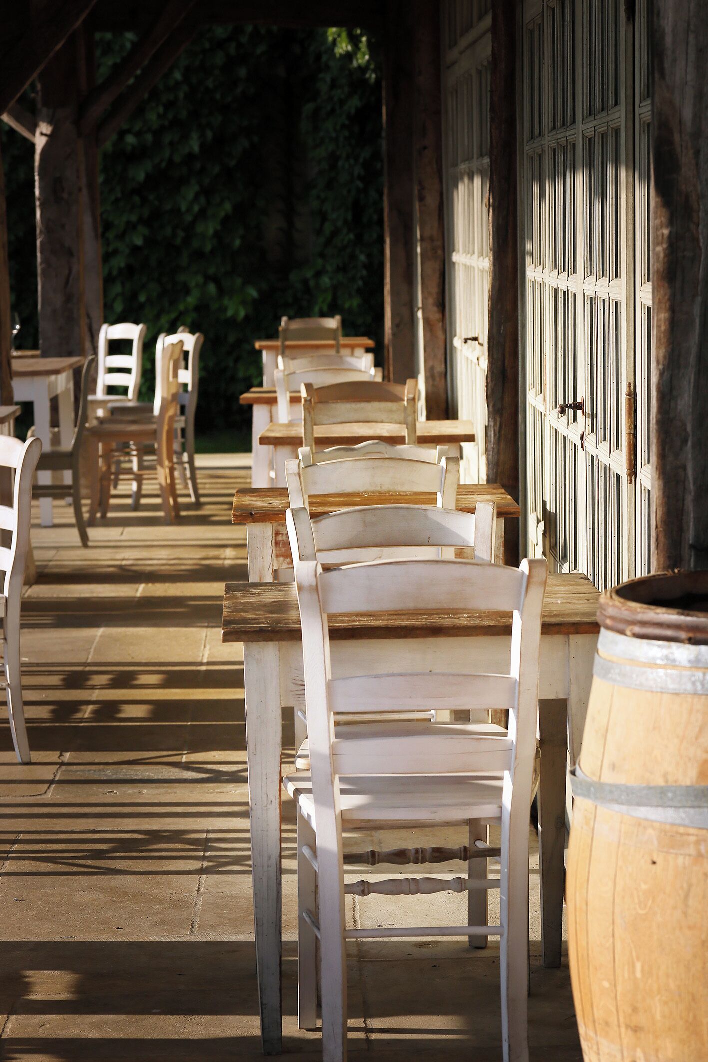 A row of white wooden chairs and tables under a covered area with a wooden barrel.